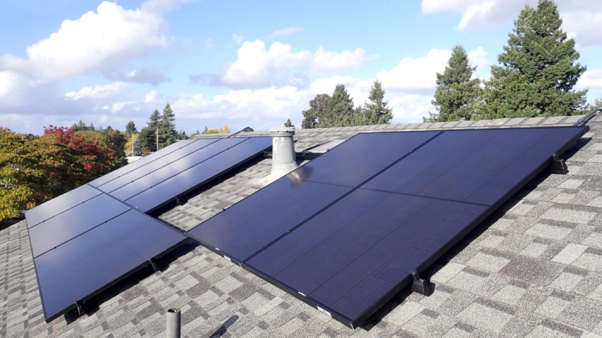 A grey asphalt tile roof with solar panels that reflect a bright blue sky with clouds.