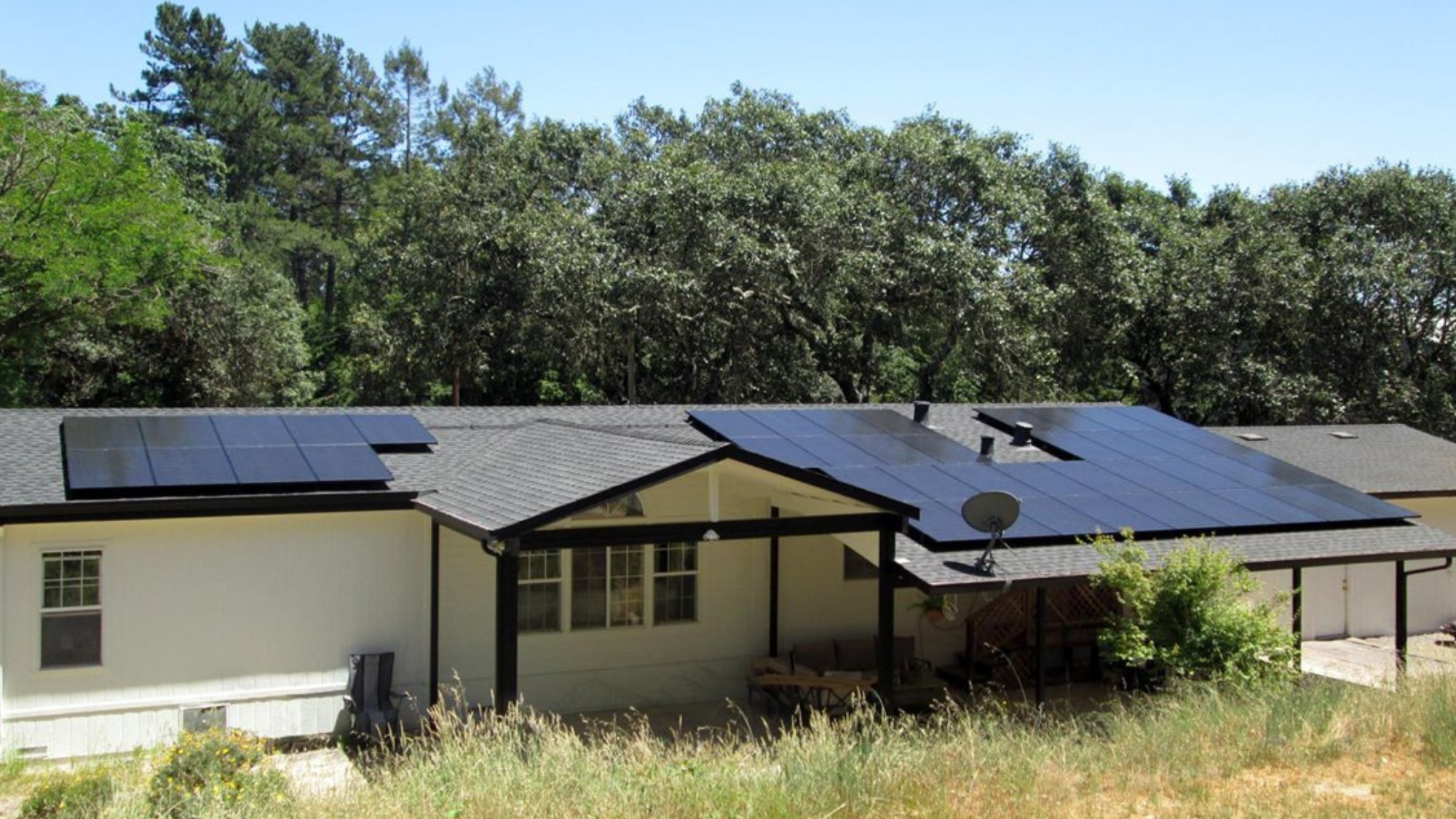 A white home with black trim with solar panels installed on the roof near oak trees.